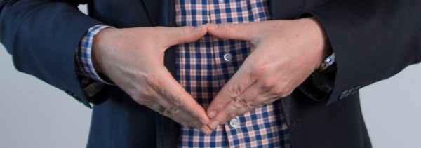 Der Bildausschnitt zeigt Mann im blauen Jacket und karietem Hemd, welche auf Höhe von Bauch gezeigt wird. Die Hände formen die typische Merkel-Raute.